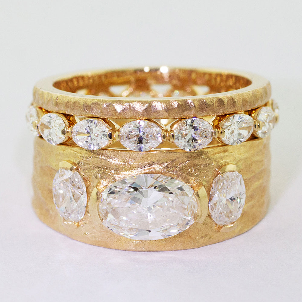 Custom 14k yellow gold engagement ring and wedding band set. The engagement ring features a bezel-set long oval diamond set in an east-west position. Adorning the shank are an additional two oval diamonds, intricate hammer texture, and a flat matte finish. The matching 14k yellow gold diamond eternity wedding band boasts fifteen bezel-set diamonds set in an east-west direction.