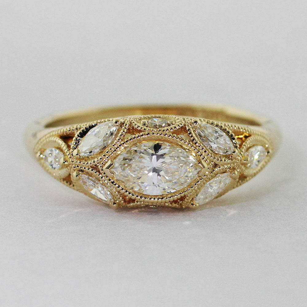 14k yellow gold Art Deco ring with a marquise-cut center diamond set in a chevron setting. Six heirloom diamonds are delicately prong-set along the 1/2 round low dome shank. Two diamond melee are set above and below the center diamond, a delicate milgrain encases each stone lending a vintage feel.
