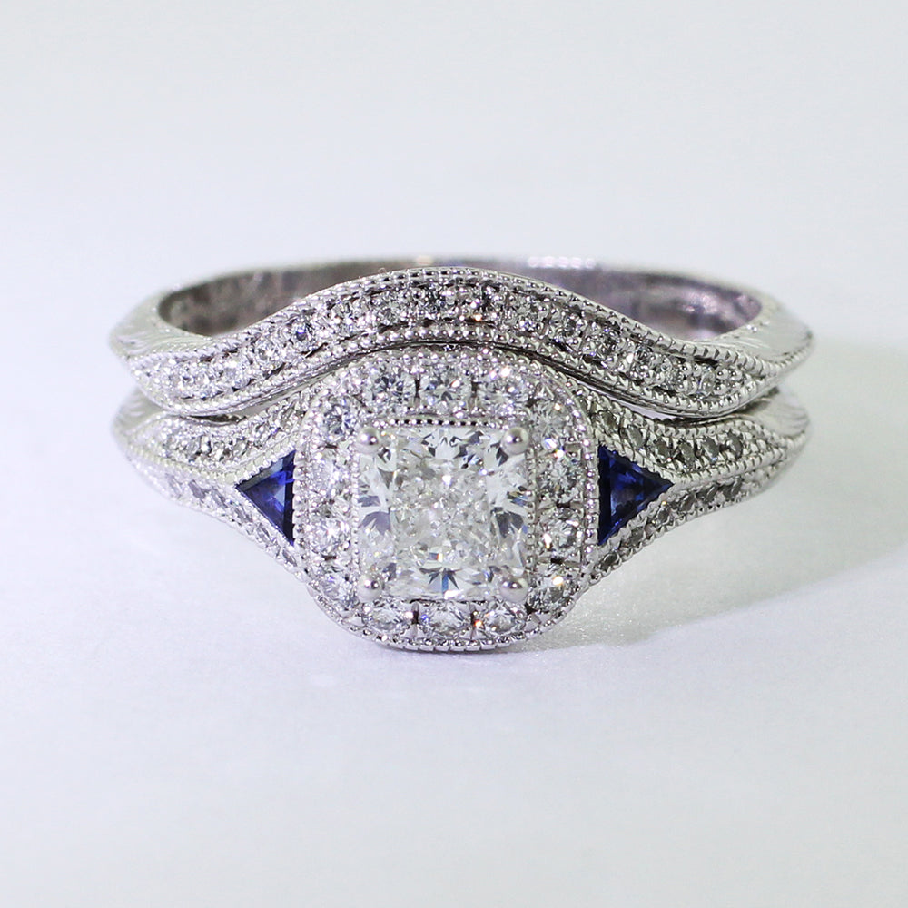 14k white gold Art Deco ring with a center cushion-cut diamond embraced by a halo of twelve 1.5mm bead-set diamonds. Two triangle sapphires add a vibrant contrast. The 14k white gold shadow band features a band of bead-set diamond melee surrounded by an engraved wheat leaf pattern and milgrain..