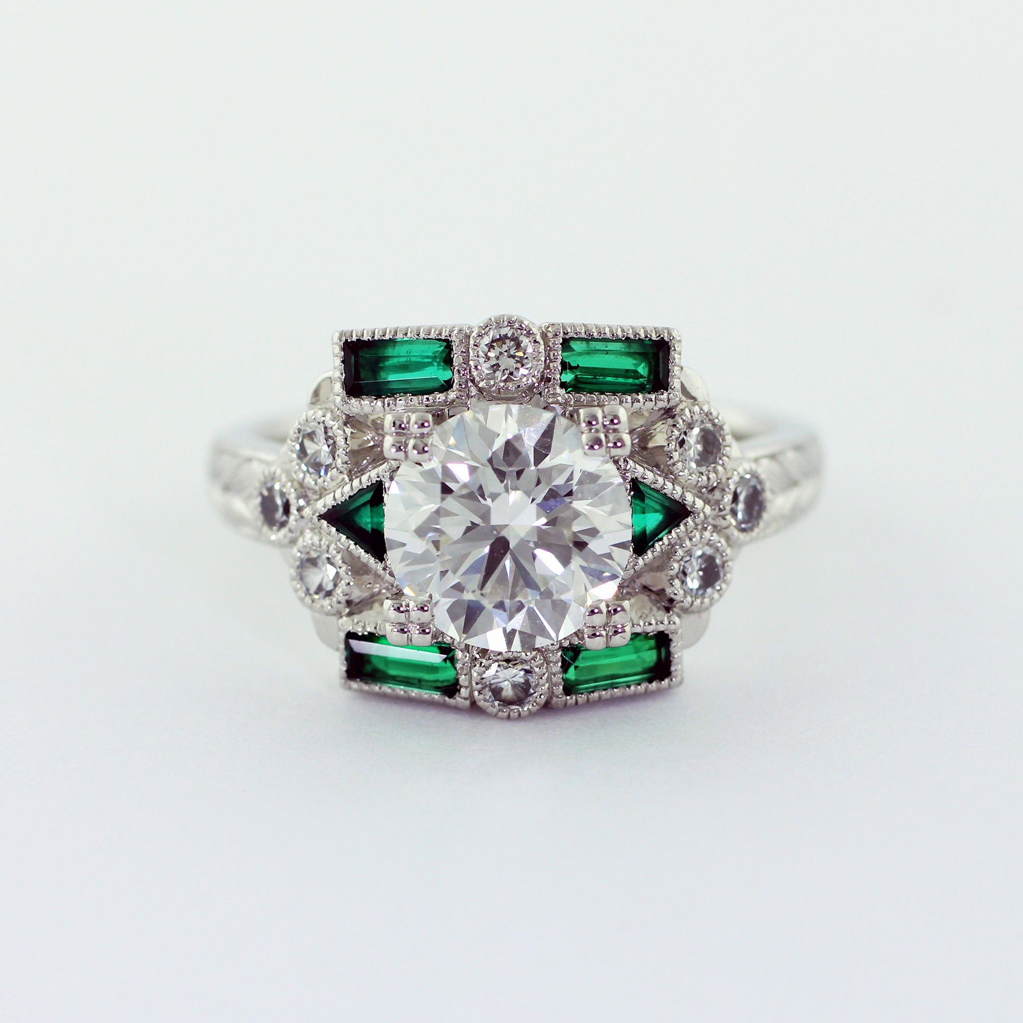 Custom Art Deco platinum ring with a prong-set 2.01ct round brilliant center diamond. The flat top shank features two triangular green emeralds, four baguette green emeralds, eight 2.2mm diamond melee, milgrain detailing, and an engraved wheat leaf pattern, lending a touch of vintage charm.