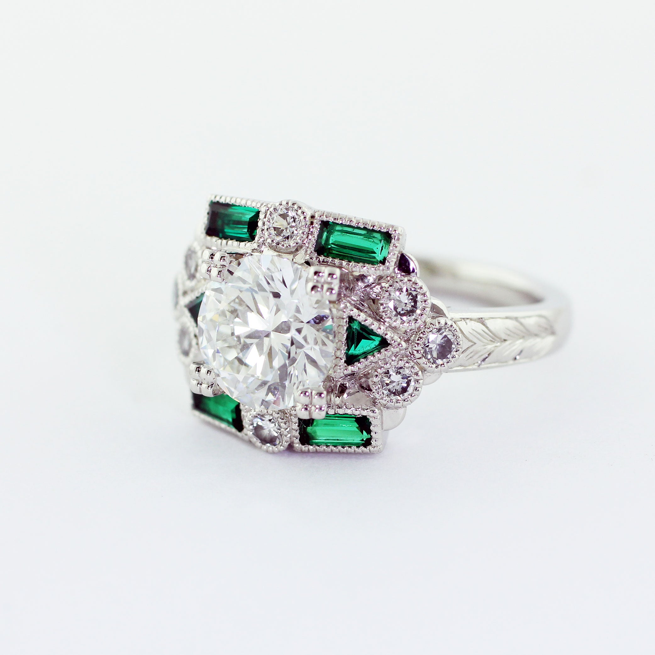 Custom Art Deco platinum ring with a prong-set 2.01ct round brilliant center diamond. The flat top shank features two triangular green emeralds, four baguette green emeralds, eight 2.2mm diamond melee, milgrain detailing, and an engraved wheat leaf pattern, lending a touch of vintage charm.