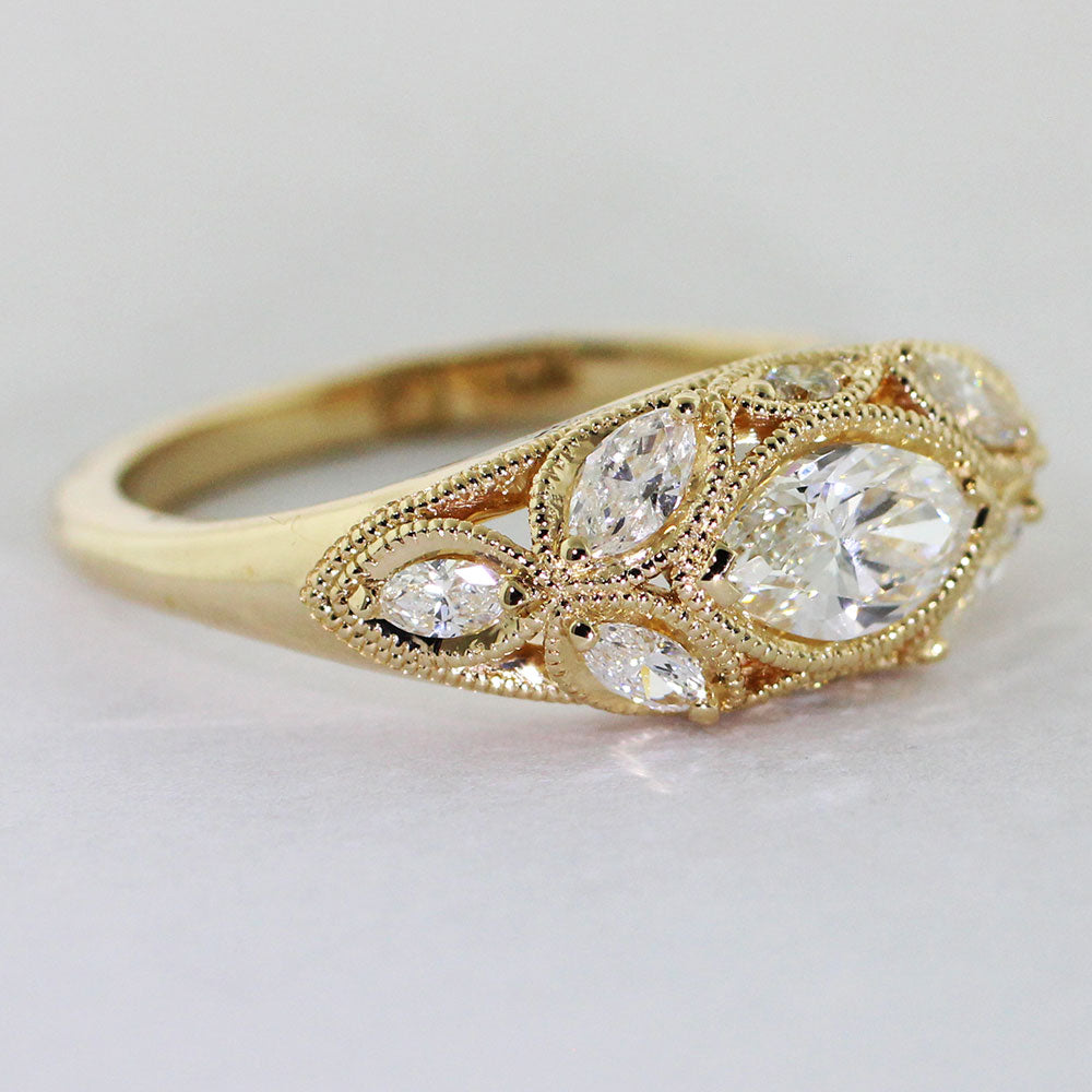 14k yellow gold Art Deco ring with a marquise-cut center diamond set in a chevron setting. Six heirloom diamonds are delicately prong-set along the 1/2 round low dome shank. Two diamond melee are set above and below the center diamond, a delicate milgrain encases each stone lending a vintage feel.