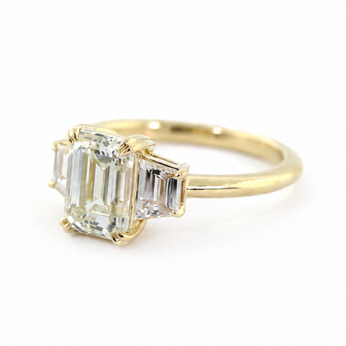 Clara engagement ring with a 1.5 ct emerald cut diamond, trapezoid side diamonds, and 14k yellow gold band.
