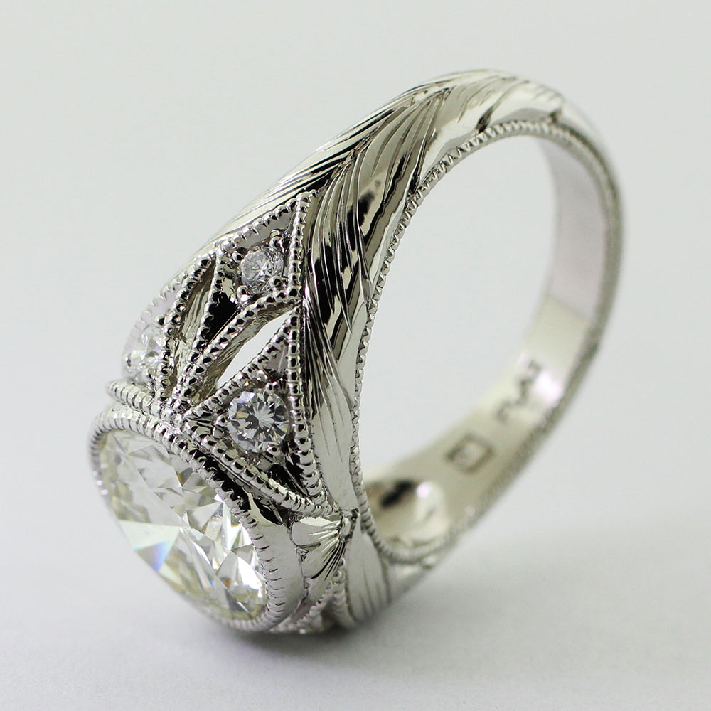 Custom platinum art deco ring featuring a round brilliant center diamond, six accent diamonds, milgrain detailing, and an engraved wheat leaf pattern.